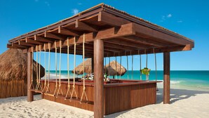 A wooden beach bar with a thatched roof, situated on a sandy beach with clear blue waters and a rocky outcrop in the background.