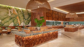 Un amplio vestíbulo de hotel con una isla central que ofrece un bufé, rodeado de zonas de estar y un gran mural en la pared.