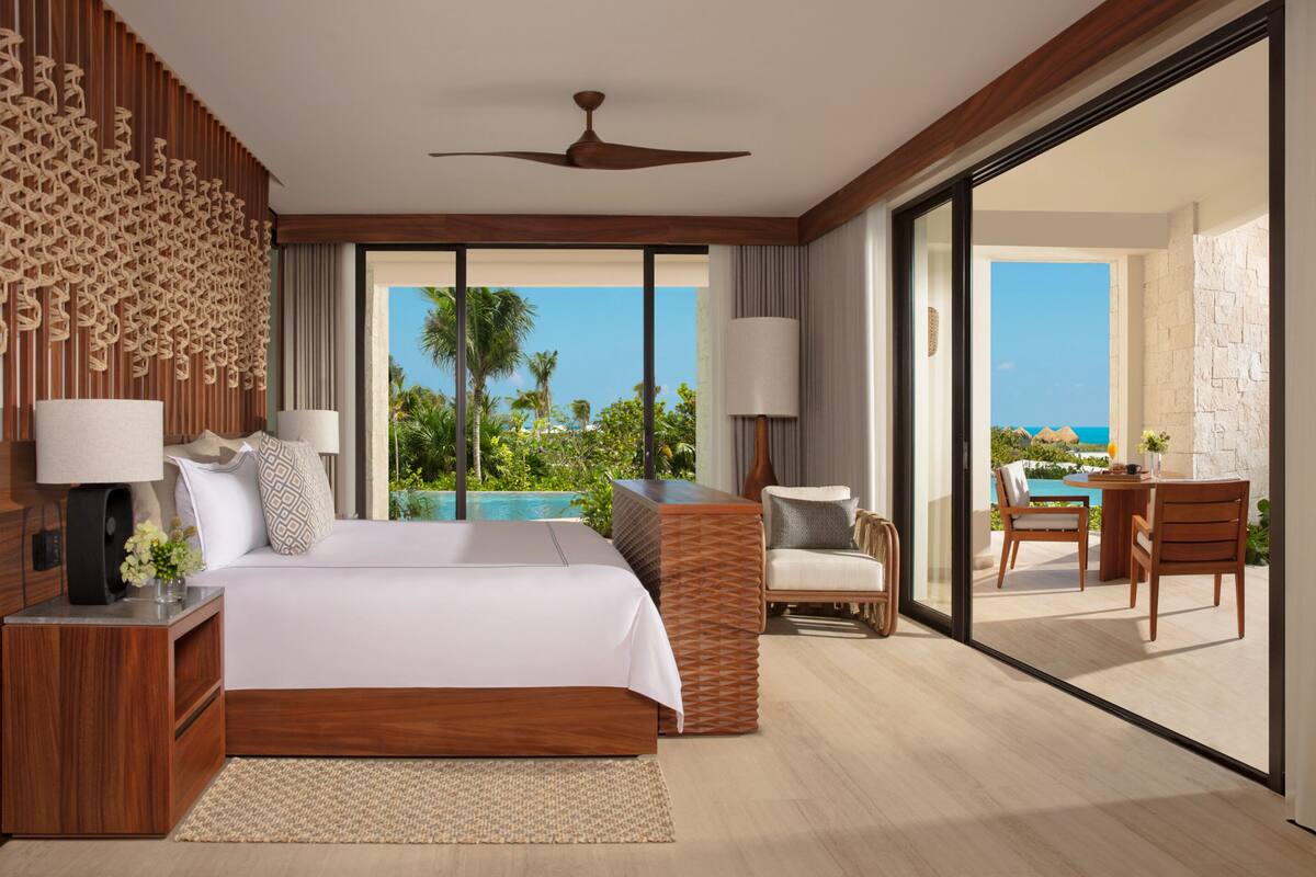 A modern hotel bedroom with a large bed, a view of the ocean, and a balcony with a pool.