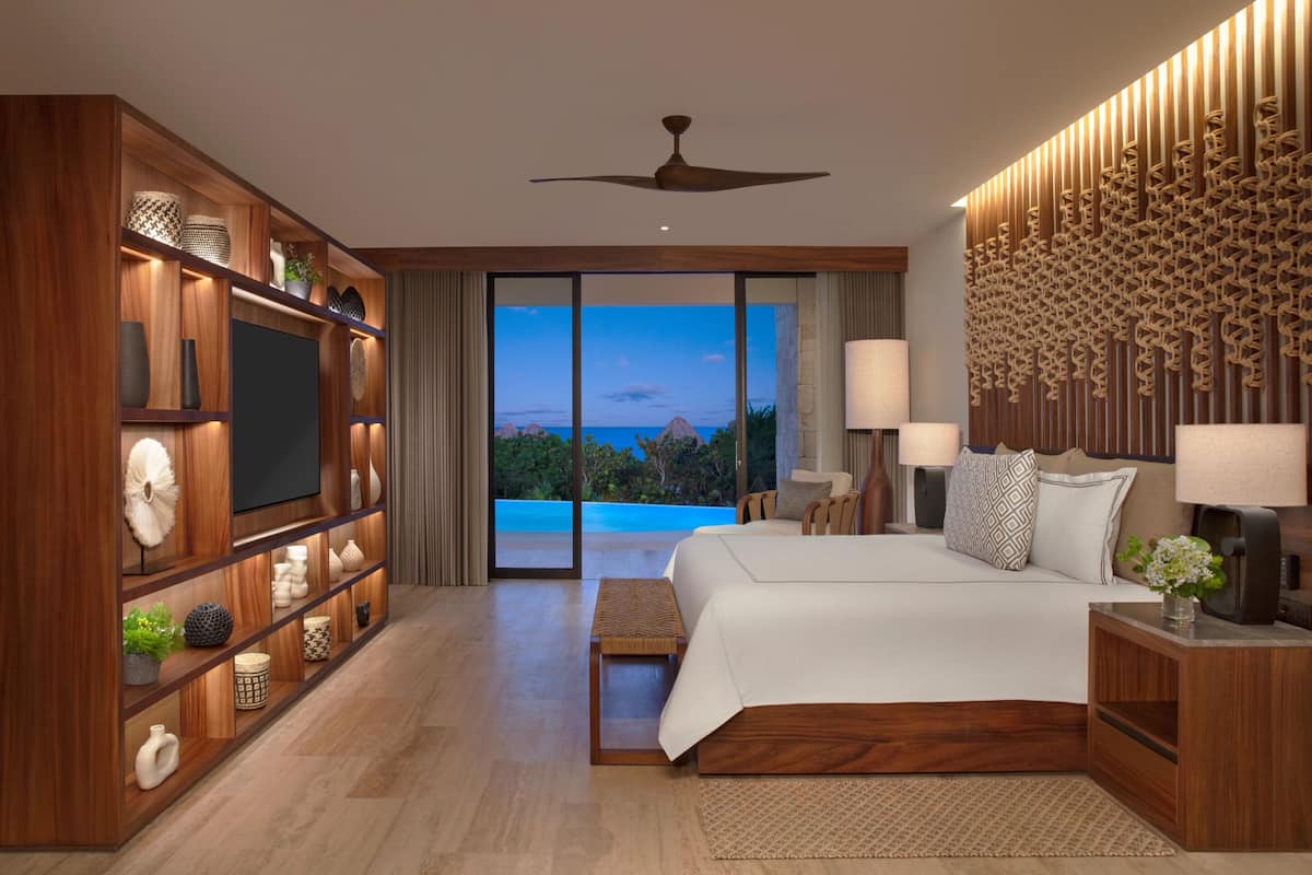 A modern hotel room with a large bed, a television, a wooden shelving unit with decorative items, a ceiling fan, and a view of the ocean.