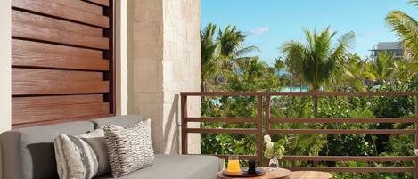 A balcony with a wooden sofa, a small table with a drink, and lush greenery in the background.