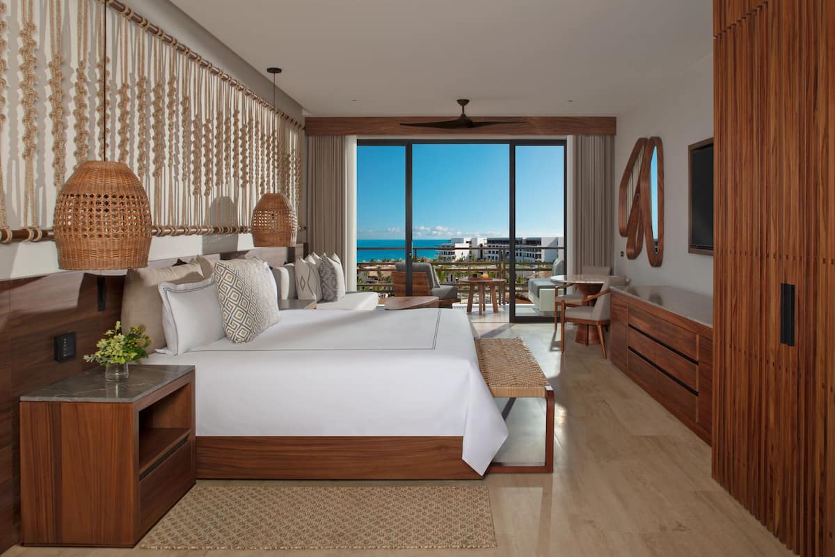 A modern hotel room with a large bed, wooden furniture, a balcony with a view of the sea, and a ceiling fan.