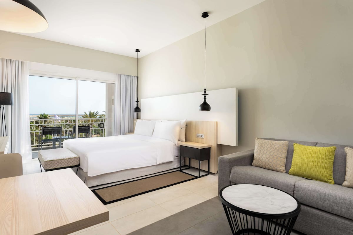 A modern hotel room with a bed, a sofa, a small table, and a balcony with a view.