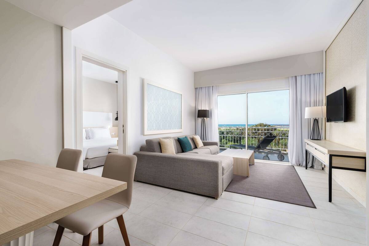 A modern hotel room with a sofa, a dining table, a TV, and a balcony with a view.
