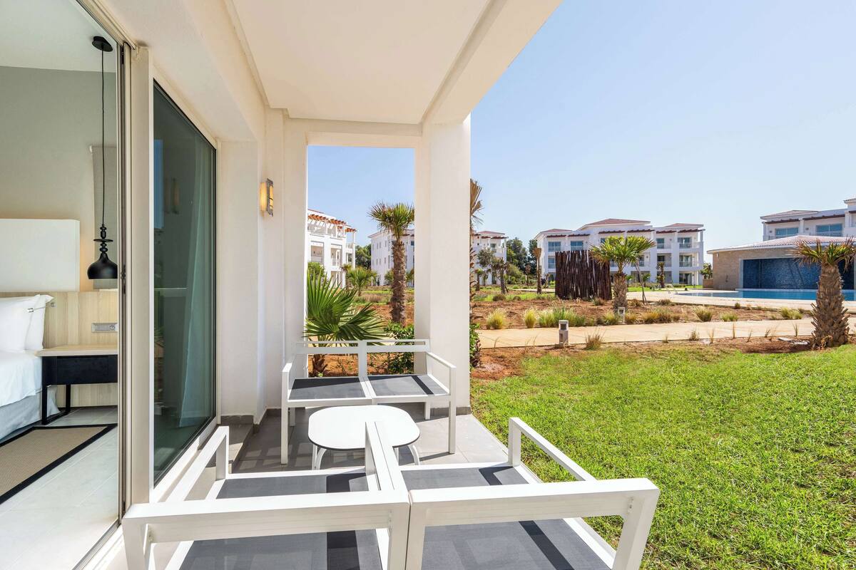A balcony with a white table and chairs, a bed, and a view of other buildings and a pool.