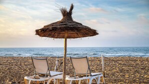 Two white lounge chairs under a thatched umbrella on a sandy beach with the ocean in the background.