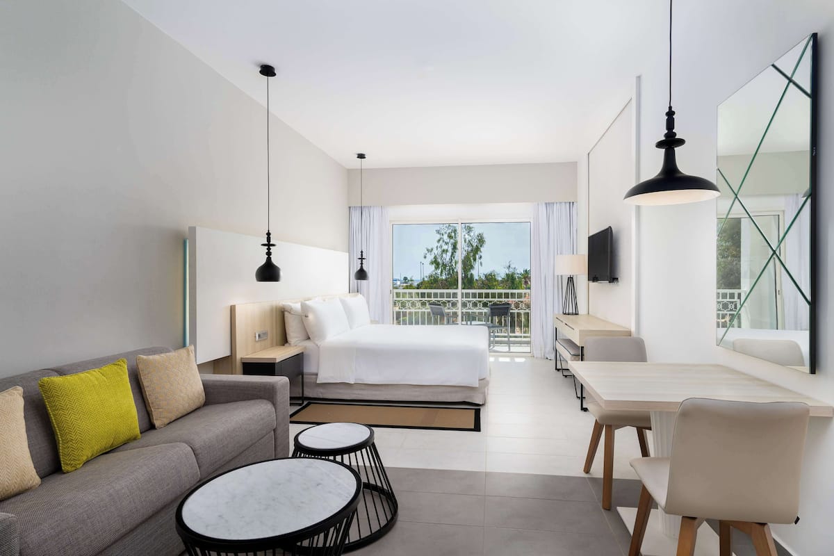 A modern hotel room with a bed, sofa, dining table, and chairs. There is a balcony with a view of greenery.