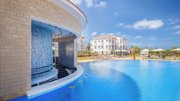A swimming pool with a curved edge, a tiled wall, and a building with a balcony in the background.