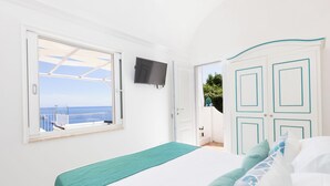 A bedroom with a bed, a TV mounted on the wall, a window with a view of the sea, and a door leading to another room. - Villa Nannina (Positano)