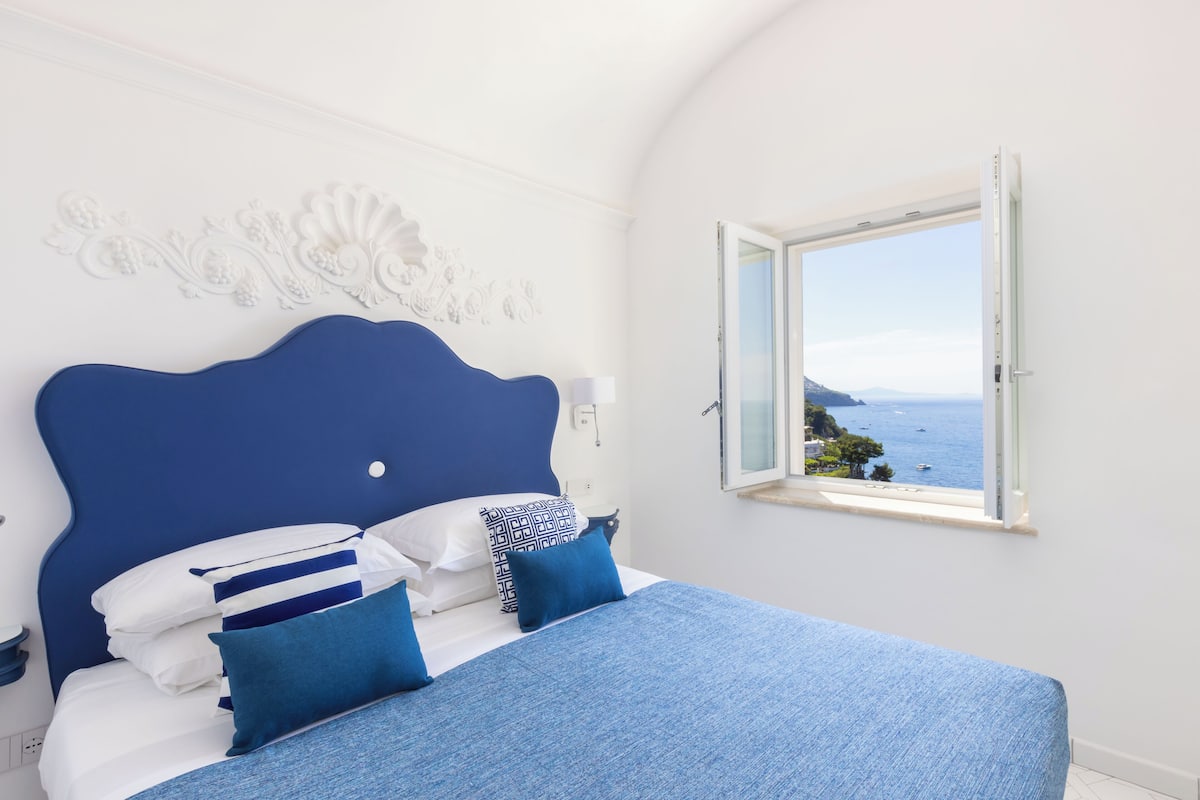 Un dormitorio con cabecera azul, paredes blancas y ventana con vistas al mar.