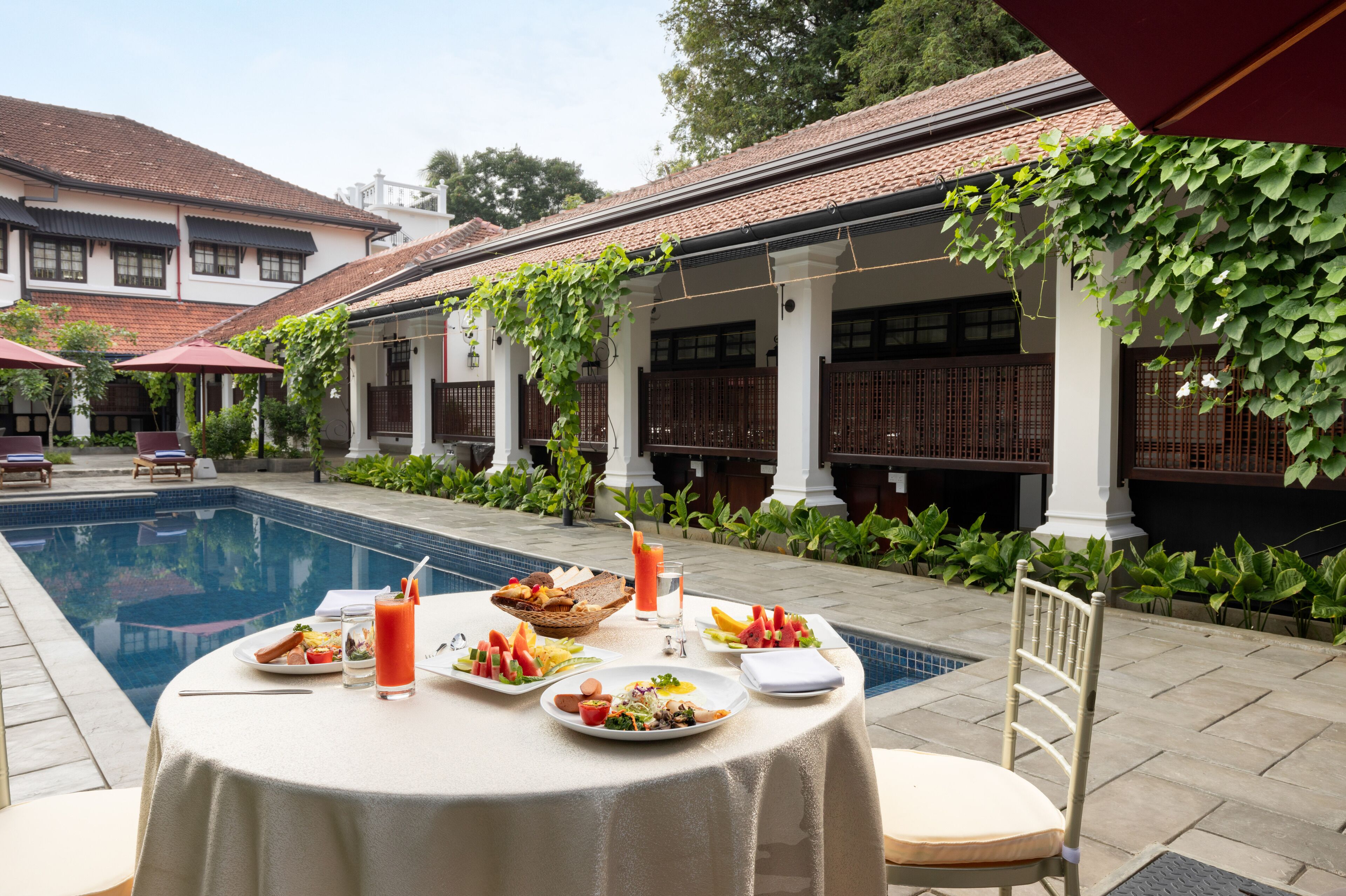 A dining table set outdoors with a pool in the background, surrounded by greenery and traditional architecture.
