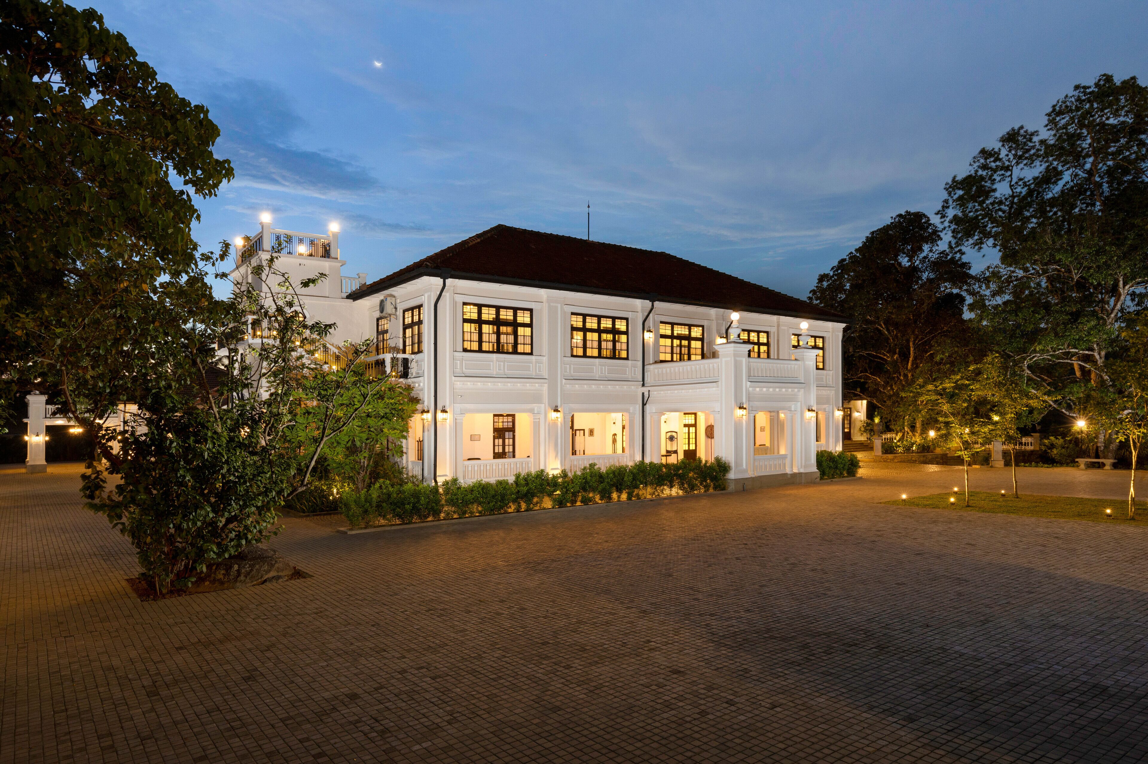 A large, white building with multiple windows and a dark roof, surrounded by trees and a paved area.