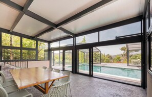 Une pièce spacieuse avec une grande table et des chaises en bois, de grandes fenêtres et une vue sur une piscine.