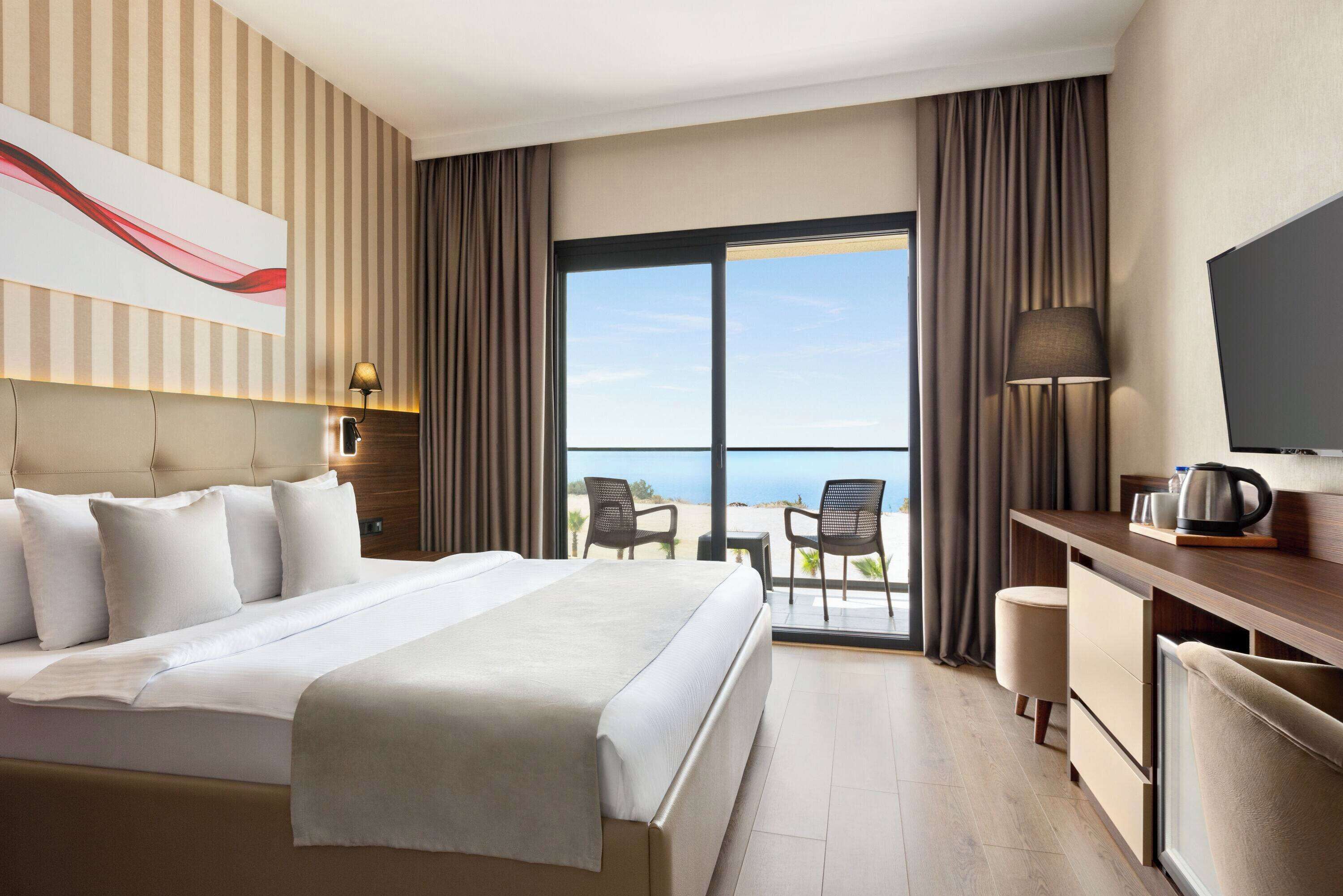 Een hotelkamer met een groot bed, een bureau met lamp, een televisie en uitzicht op het strand.