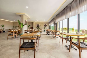 A spacious dining area with multiple tables and chairs, large windows, and potted plants.