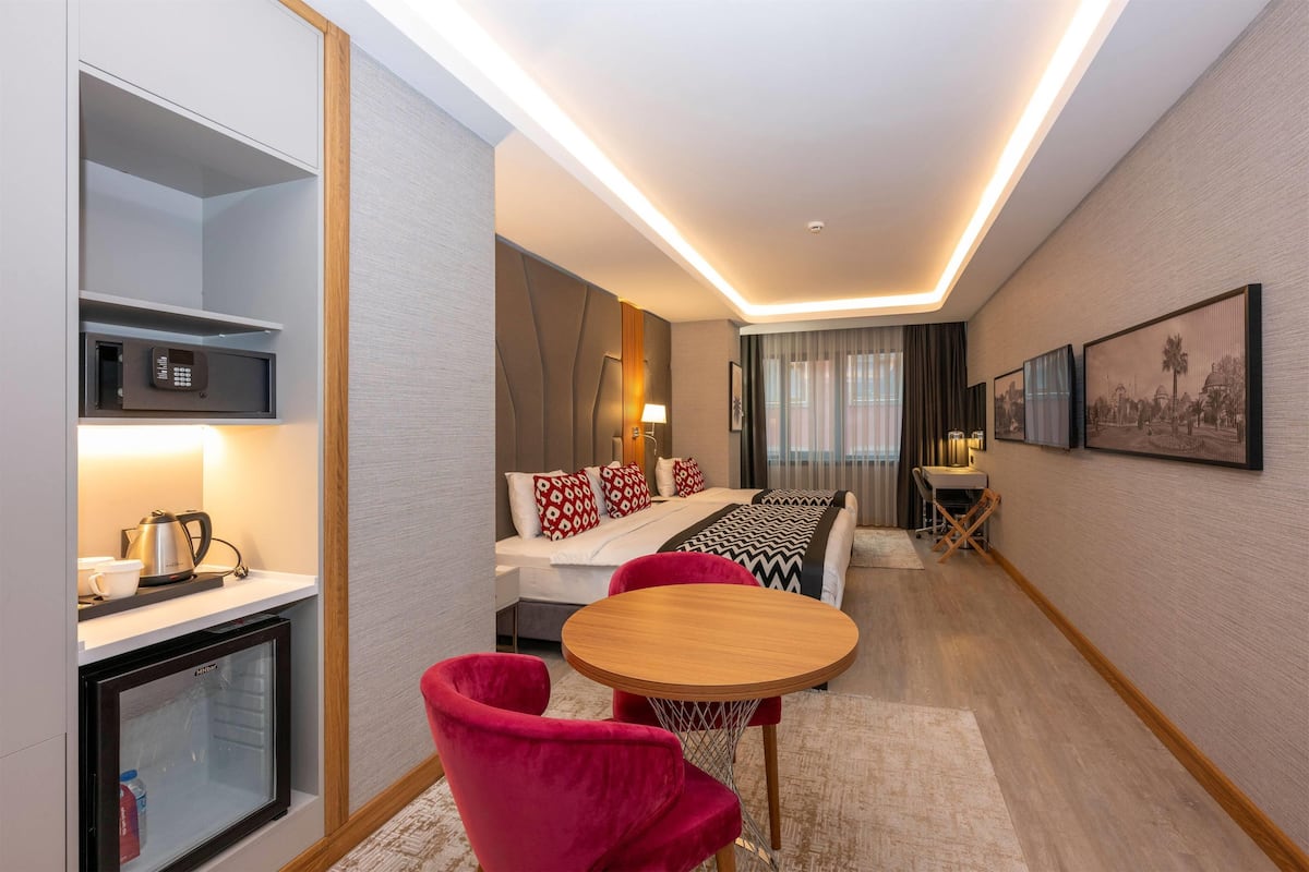A modern hotel room with a bed, a desk with a phone, a small refrigerator, and a red chair.
