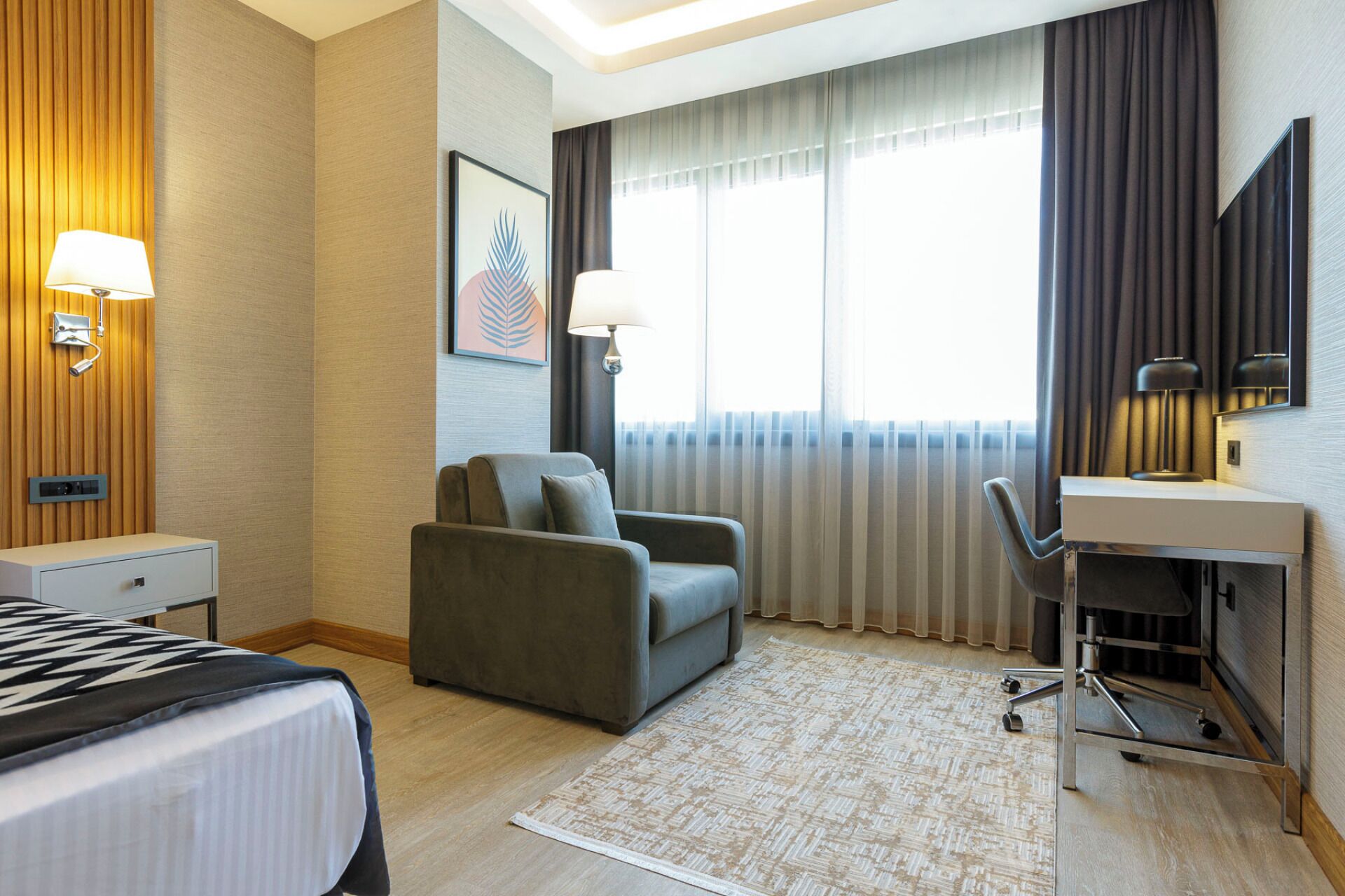 A hotel room with a bed, a desk, a chair, a lamp, a picture on the wall, and a window with curtains.