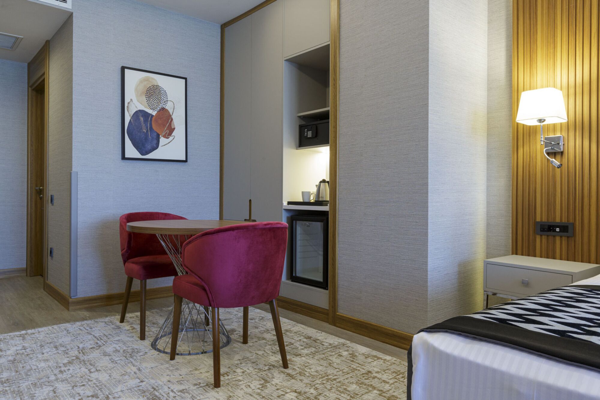 A hotel room with a bed, a desk with a chair, a kitchenette, and a wall-mounted lamp.