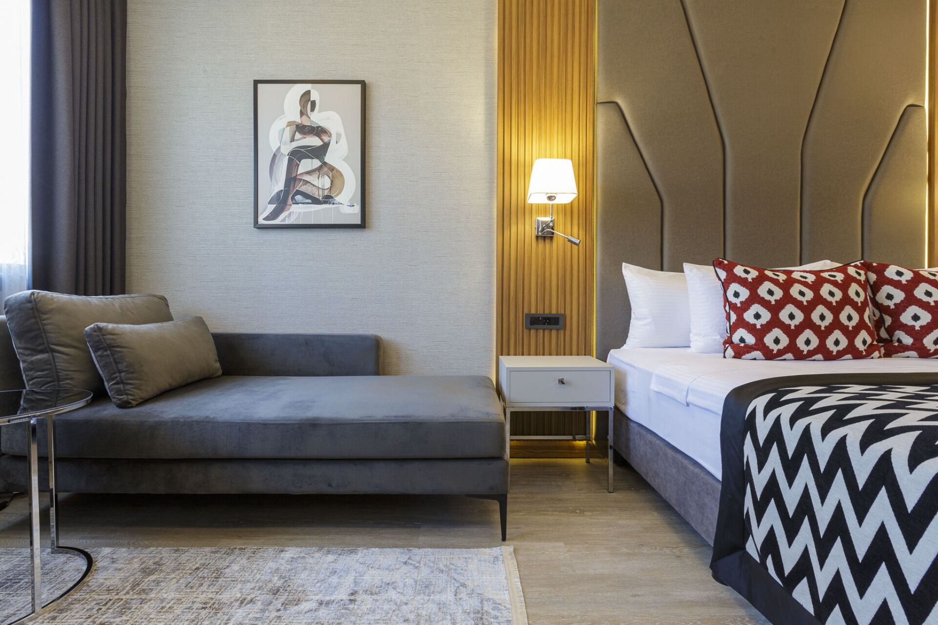 A modern hotel room with a grey sofa, a bed with patterned pillows, a nightstand, a bedside lamp, and a framed artwork on the wall.