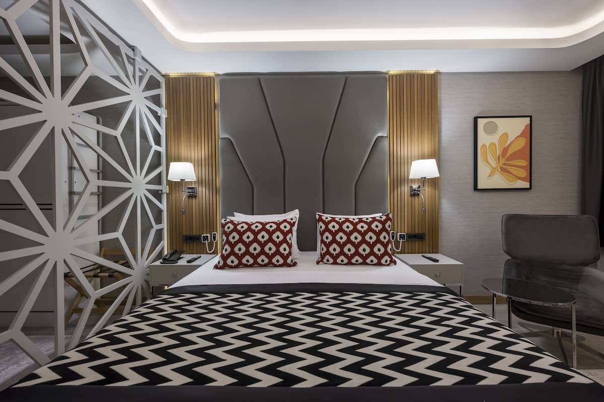 A modern hotel room with a large bed, two nightstands, a desk, a chair, and a decorative wall panel.