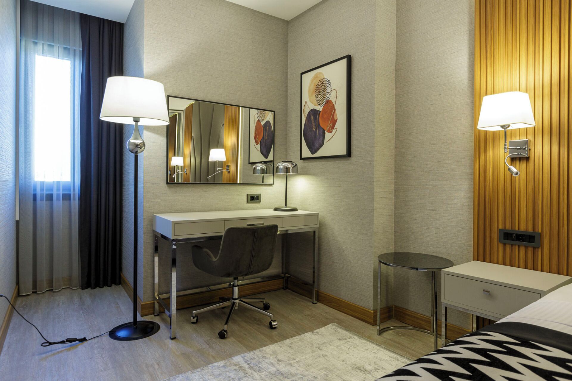 Una habitación de hotel con escritorio, silla, dos lámparas, cama y una mesita.