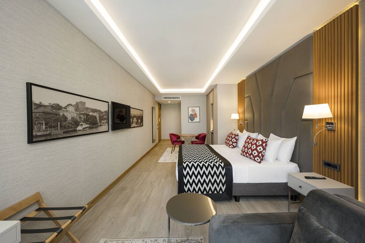 A modern hotel room with a bed, bedside tables, a desk, and a seating area with chairs.