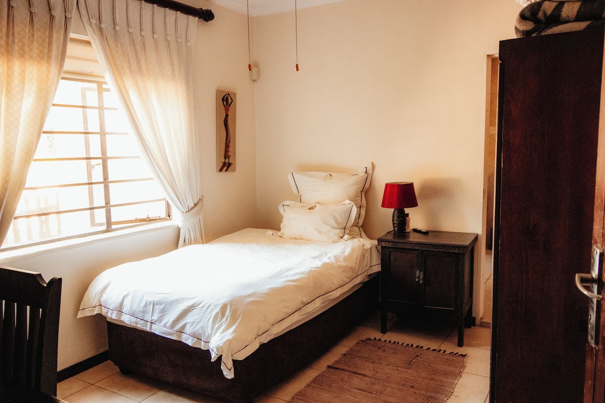 Een slaapkamer met een enkel bed, nachtkastje, stoel, houten garderobe en een raam met gordijnen.