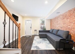 A modern living room with a grey sofa, a small black side table, and a brick accent wall. - Charm City Oasis (Baltimore)