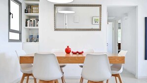 Un coin repas avec une table en bois, des chaises blanches, un vase rouge et une bibliothèque.