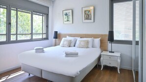 Une chambre à coucher avec un grand lit, deux tables de chevet, une commode et deux fenêtres équipées de stores.