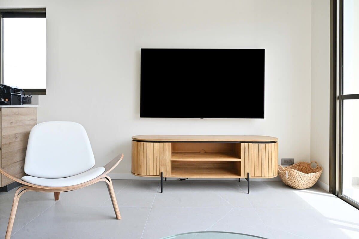 A modern living room with a flat-screen TV mounted on the wall, a white chair, a wooden console table, and a large window providing natural light.