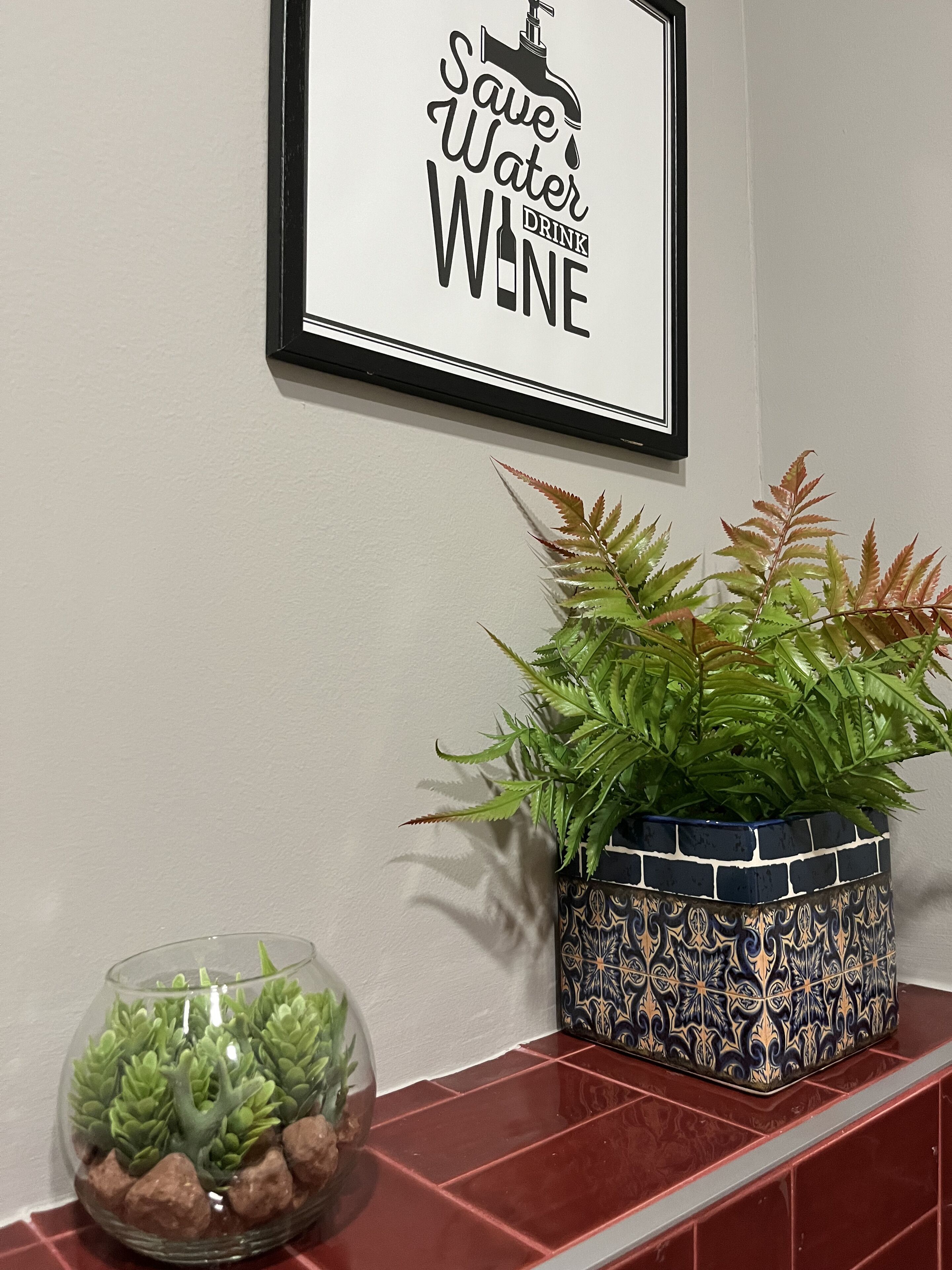 Un cartel enmarcado con el texto 'Save Water Drink Wine' y dos plantas en macetas sobre una superficie de baldosas.