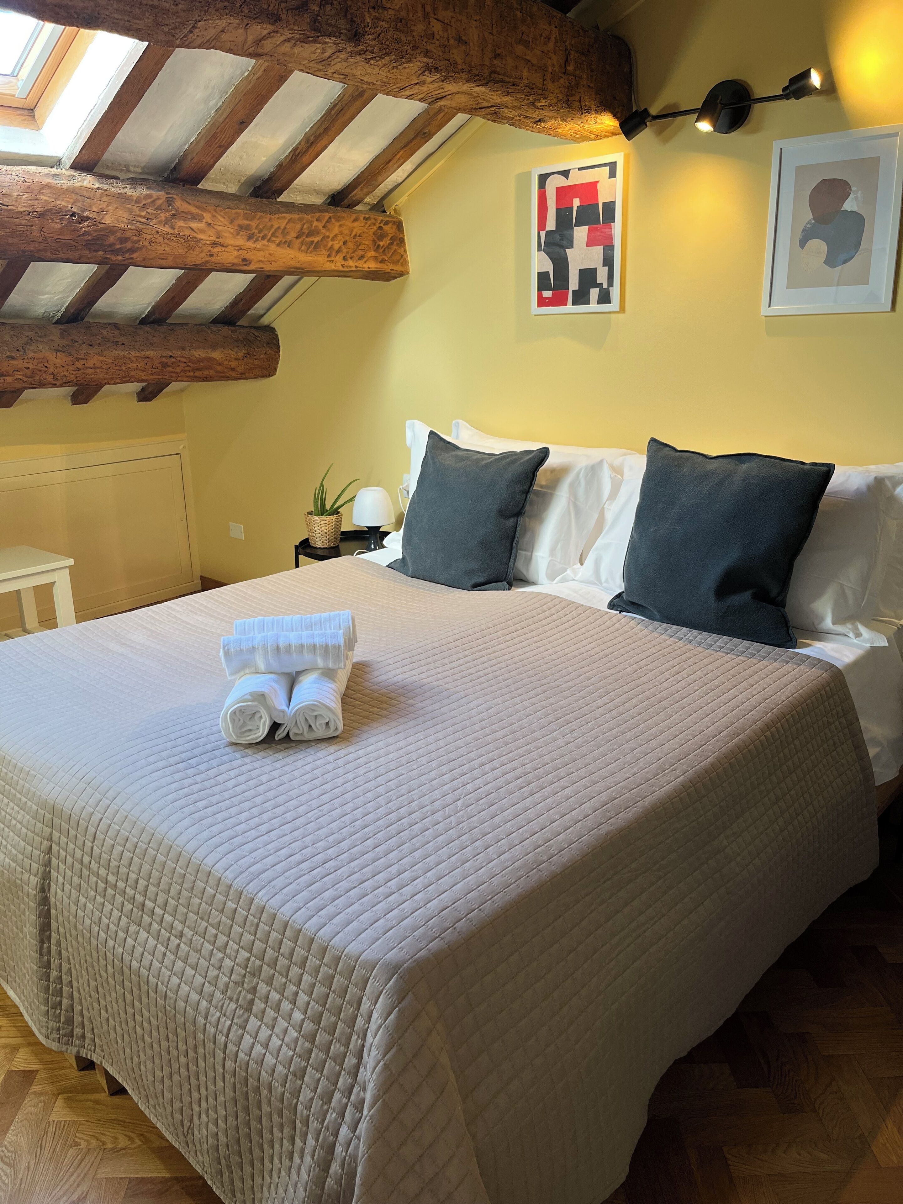 A neatly made bed with a quilted bedspread, two pillows, and rolled towels on a wooden floor.