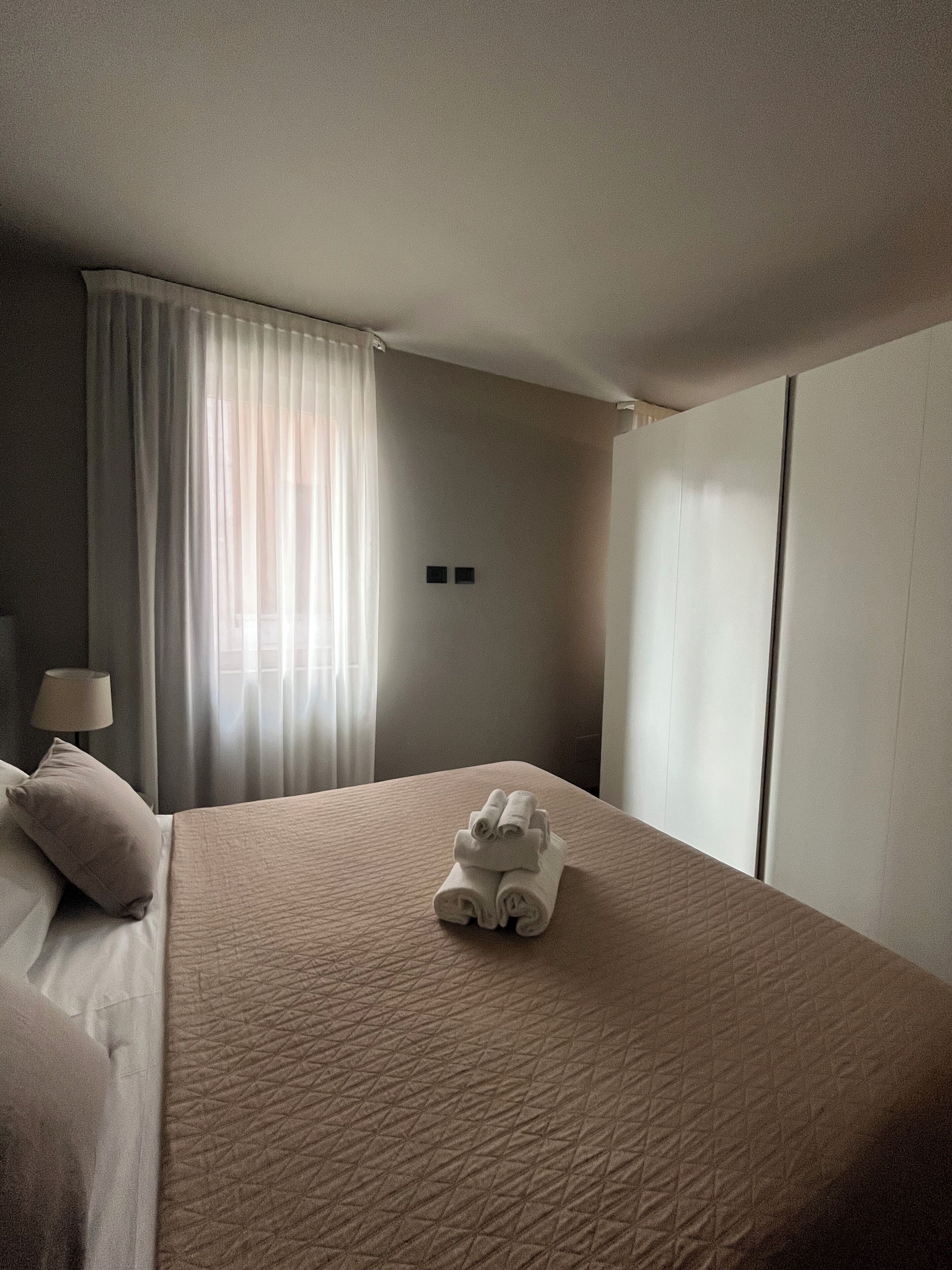 Een hotelkamer met een groot bed, een nachtkastje met een lamp en een raam met gordijnen.