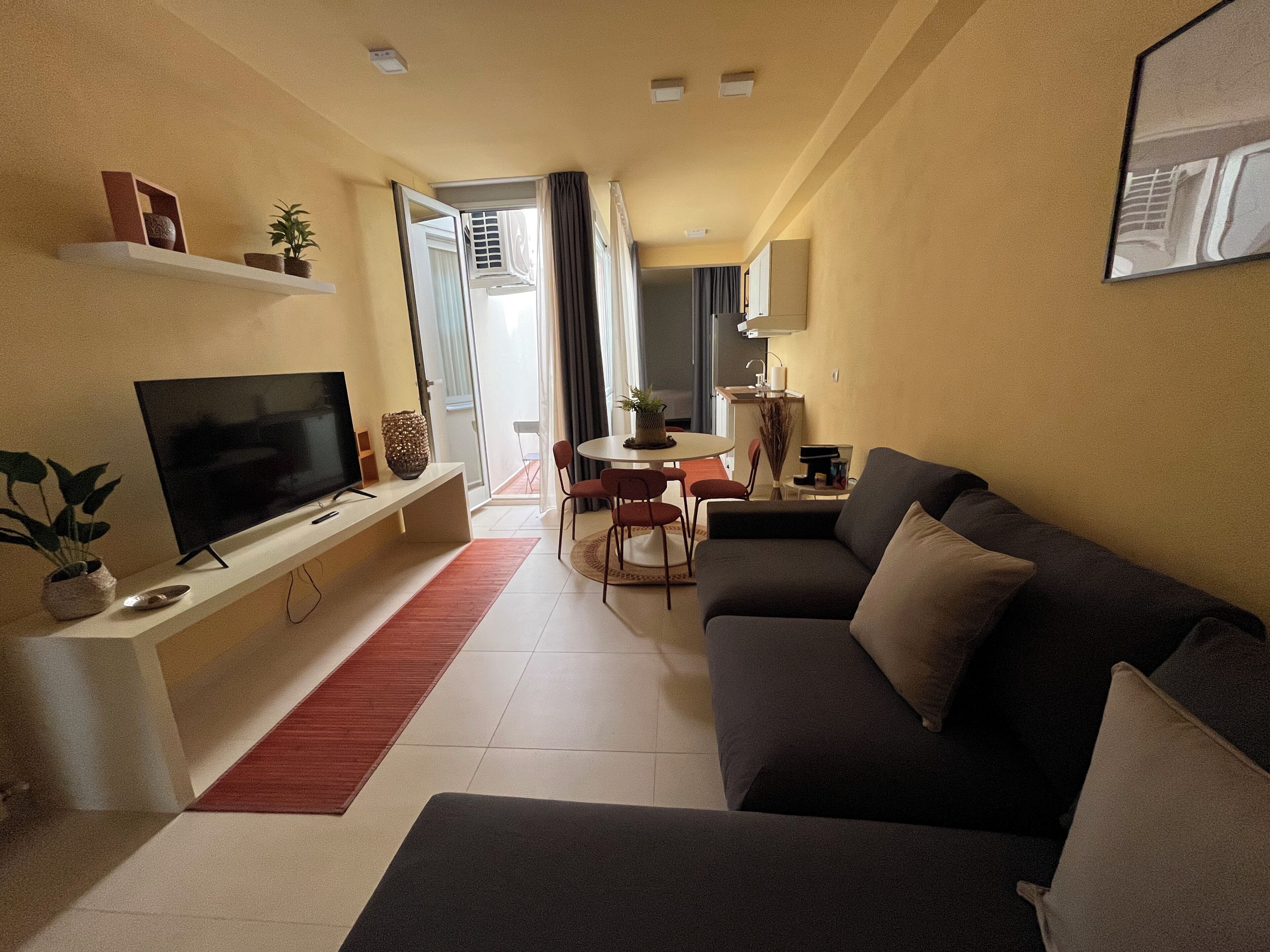 A modern living room with a flat-screen TV, a sofa, a dining table with chairs, and a kitchen area in the background.