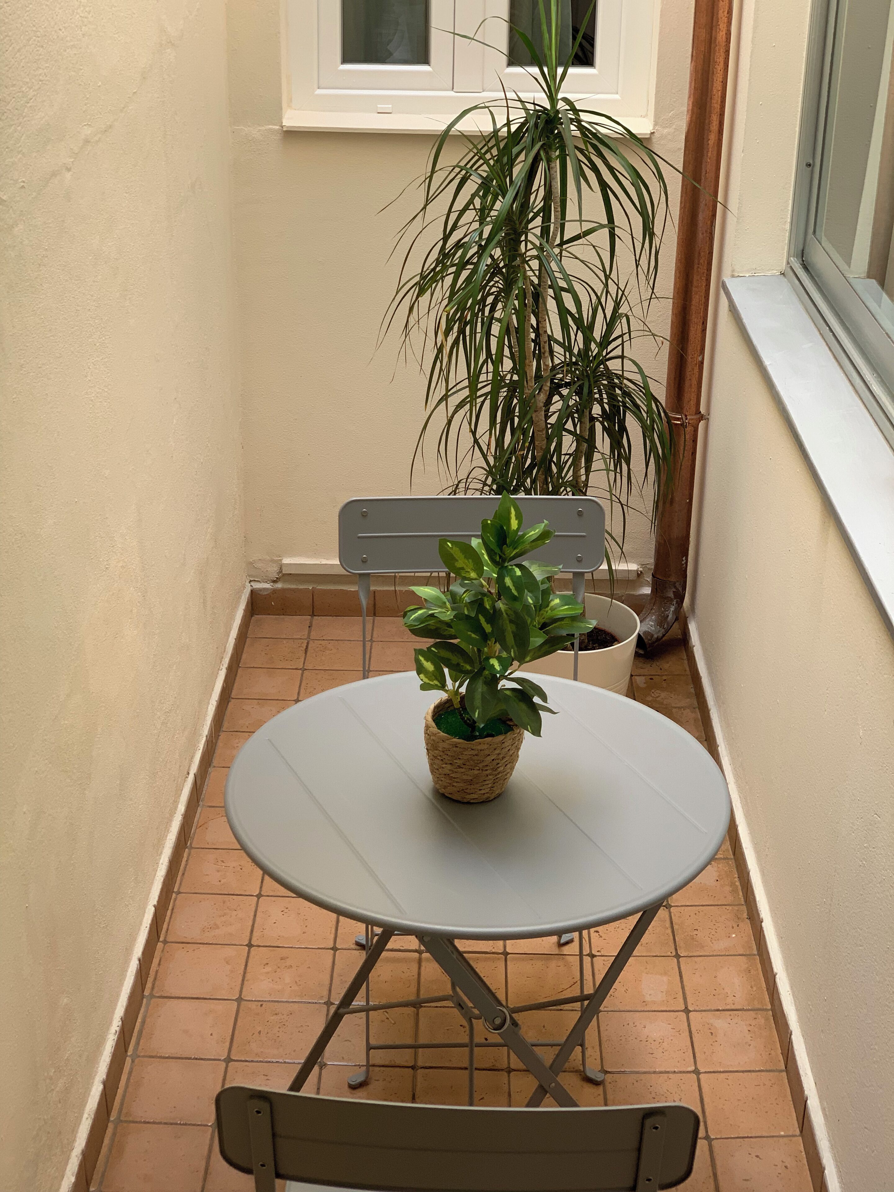 A small outdoor patio with a round table and two chairs, a potted plant, and a window with blinds.