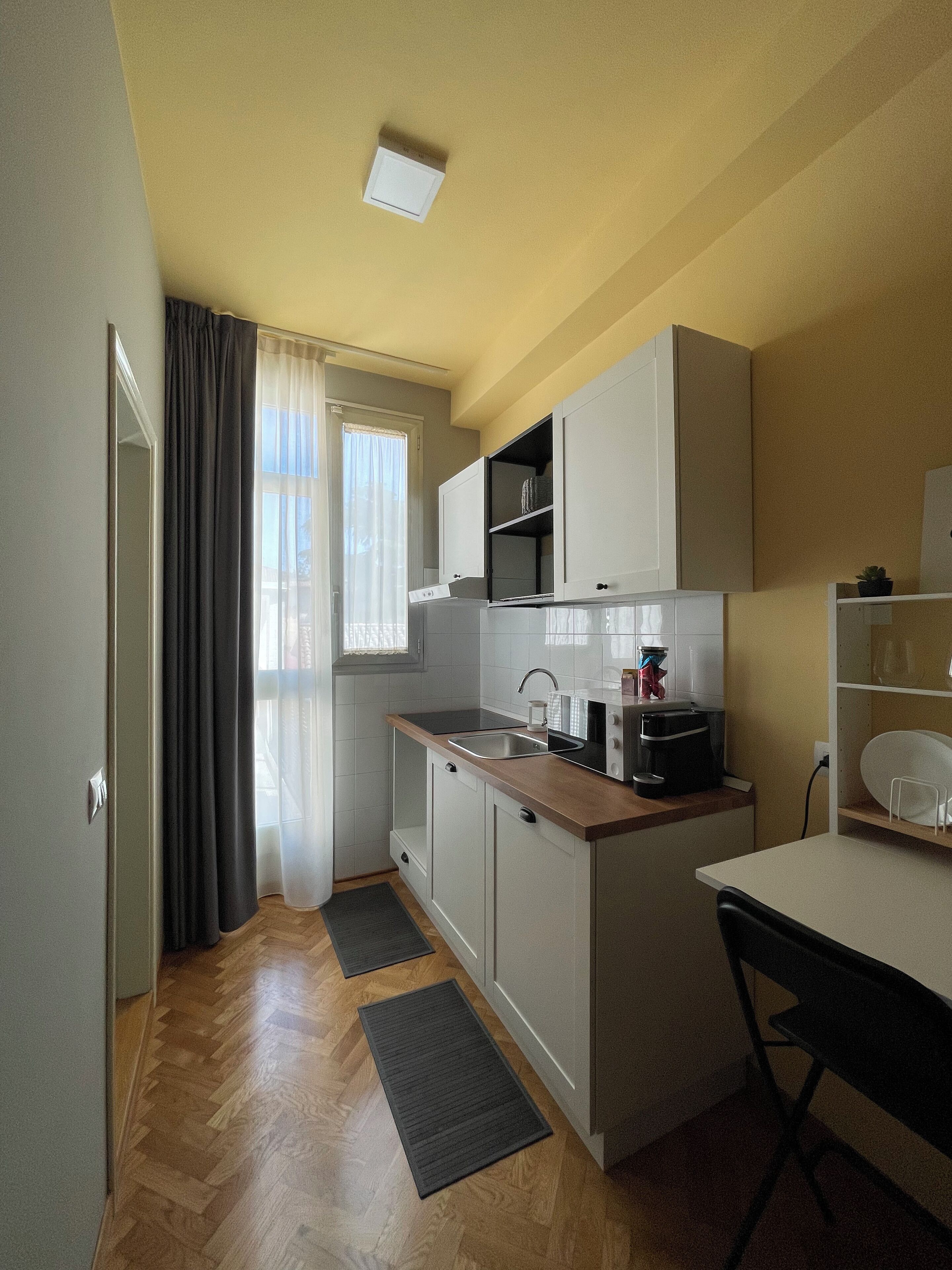 Una cuina petita amb un rentapelats, fogó i armaris, una zona de dinar reduïda amb una cadira, i una finestra amb cortines.