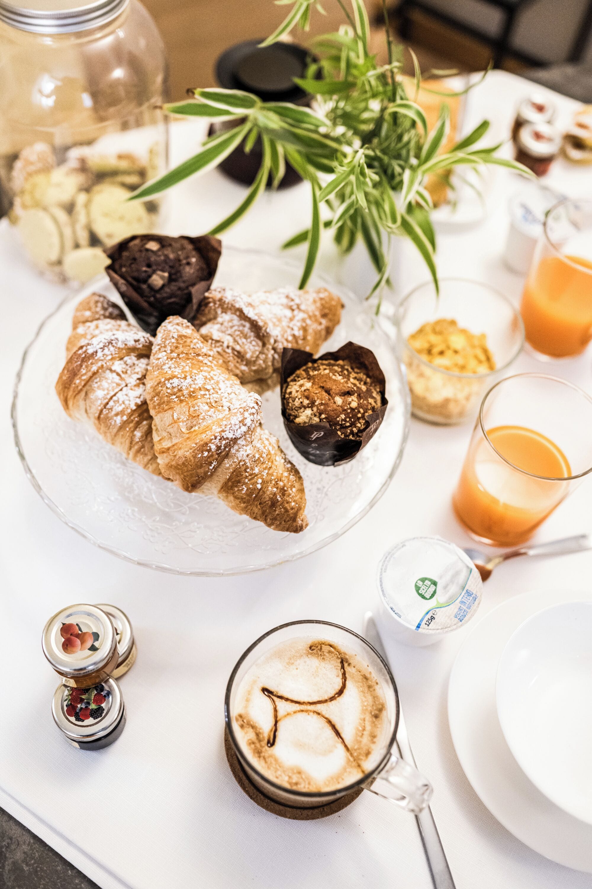 Desayuno con bollos, una taza de café y un vaso de jugo de naranja sobre una mesa blanca.