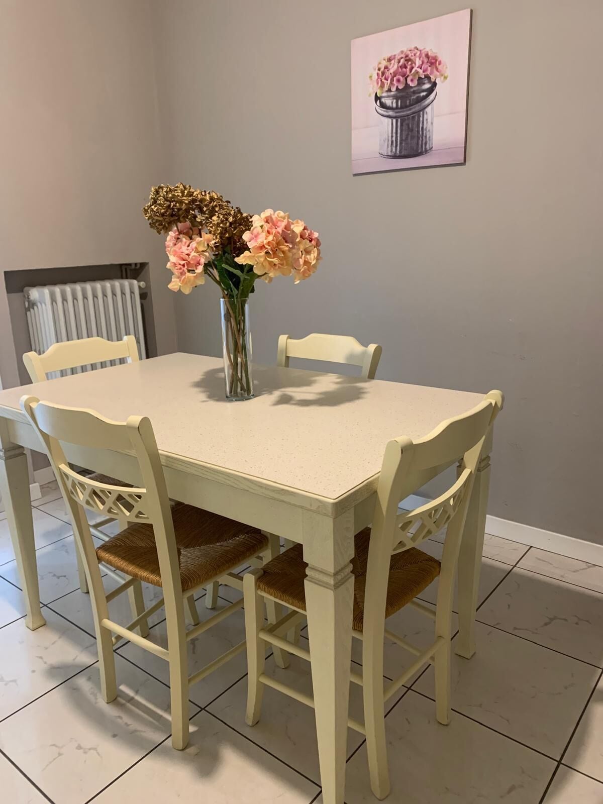 A dining table with four chairs, a vase with flowers, and a painting on the wall.