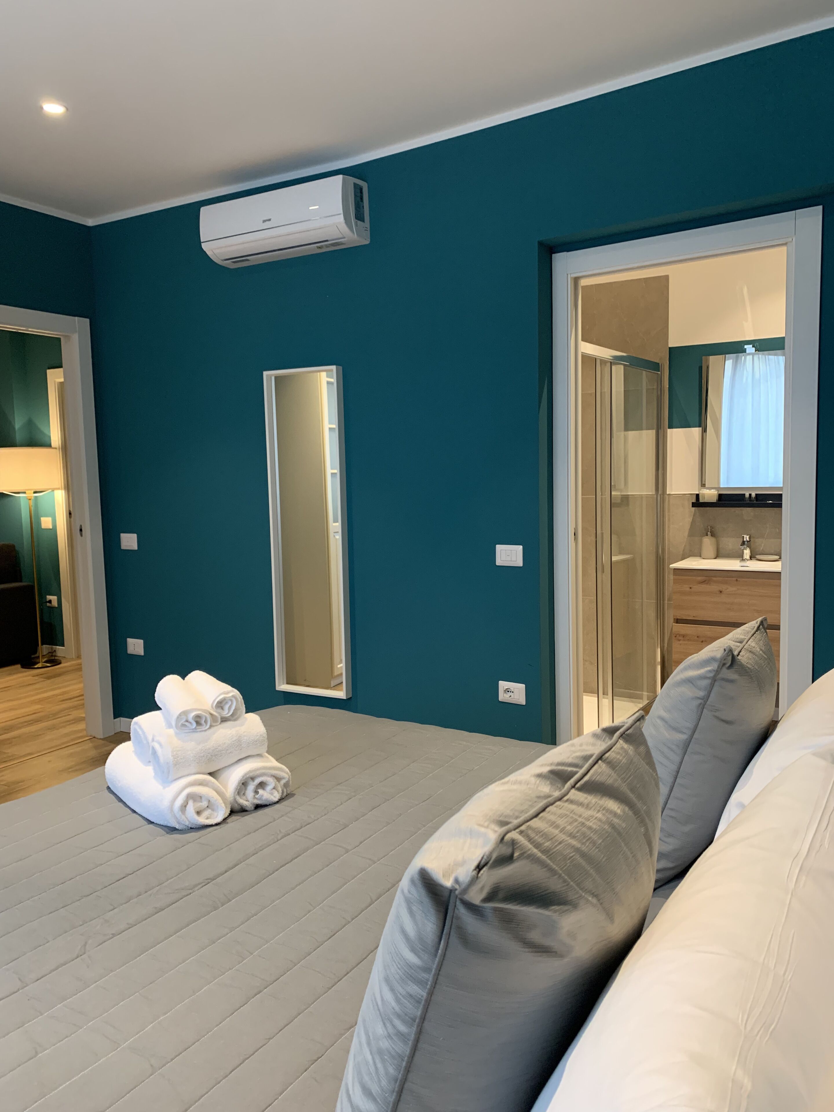 A modern hotel room with teal walls, a large mirror, a glass shower enclosure, and neatly folded towels on the floor.