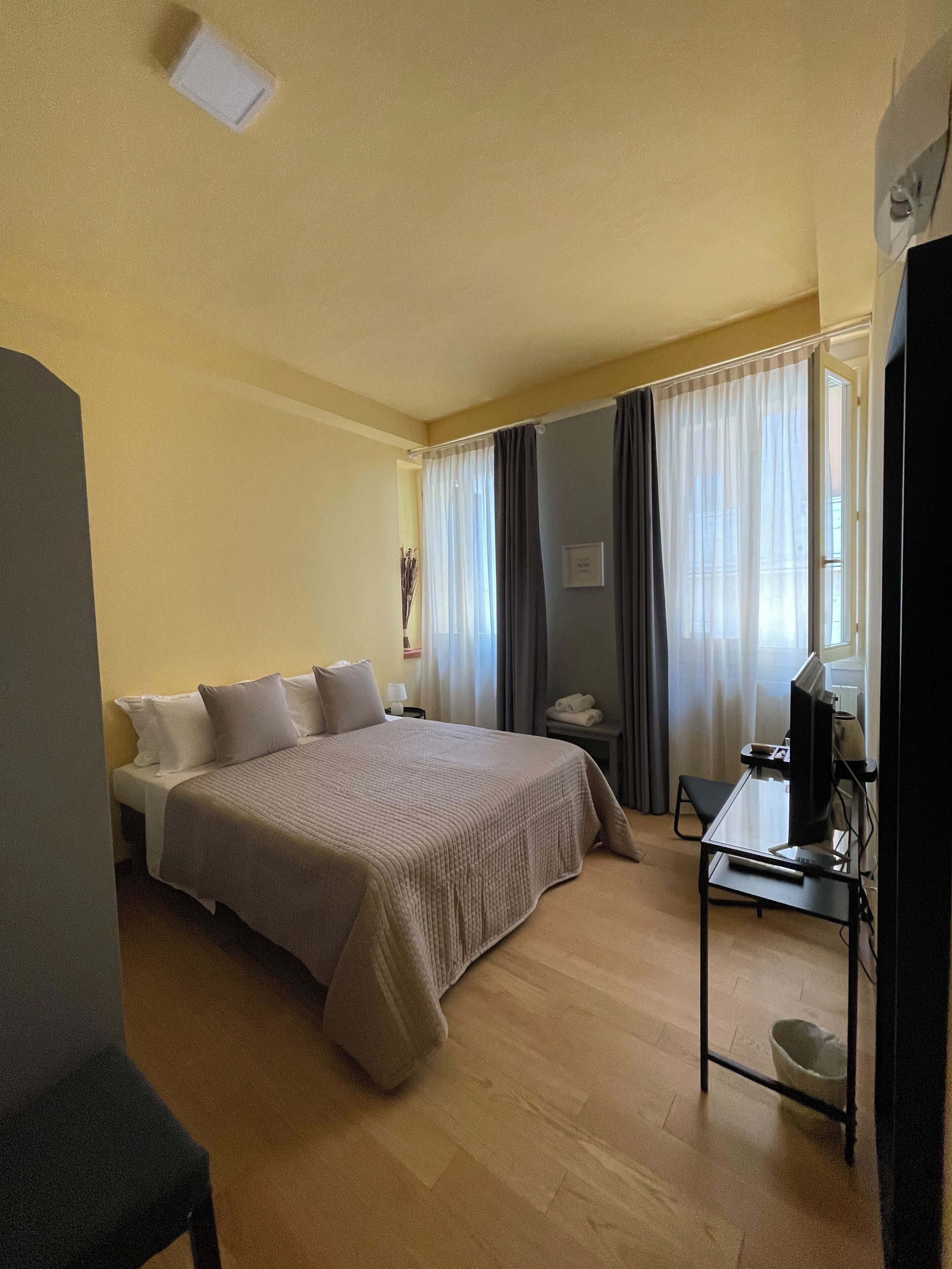 Una habitación de hotel con cama, escritorio, computadora, silla y ventana con cortinas.