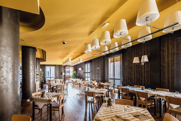 Un restaurante espacioso con techos altos, lámparas colgantes modernas y una larga zona de comedor con mesas y sillas.