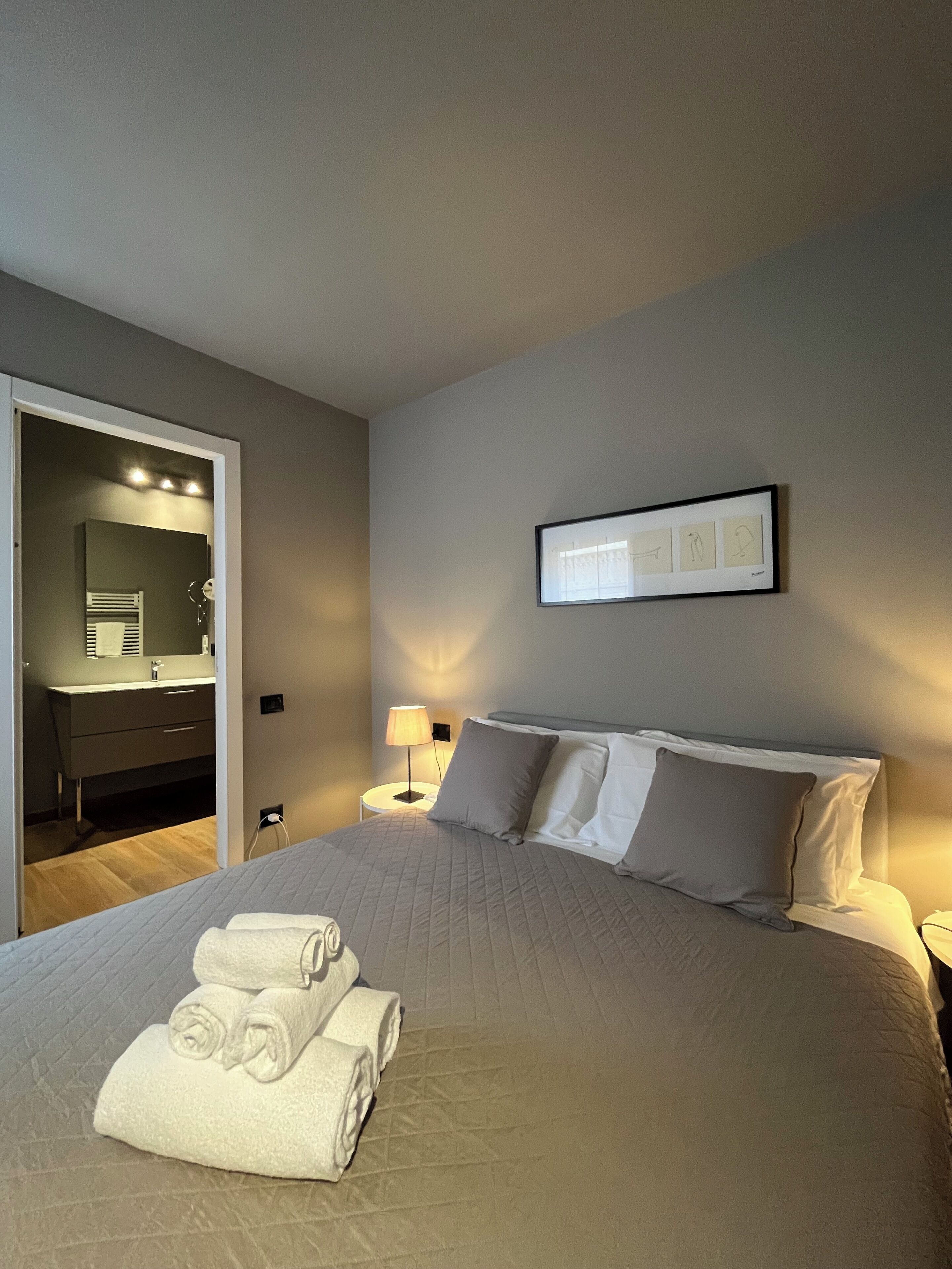 Una cama bien hecha con toallas enrolladas, una lámpara de noche y un cuadro en la pared.