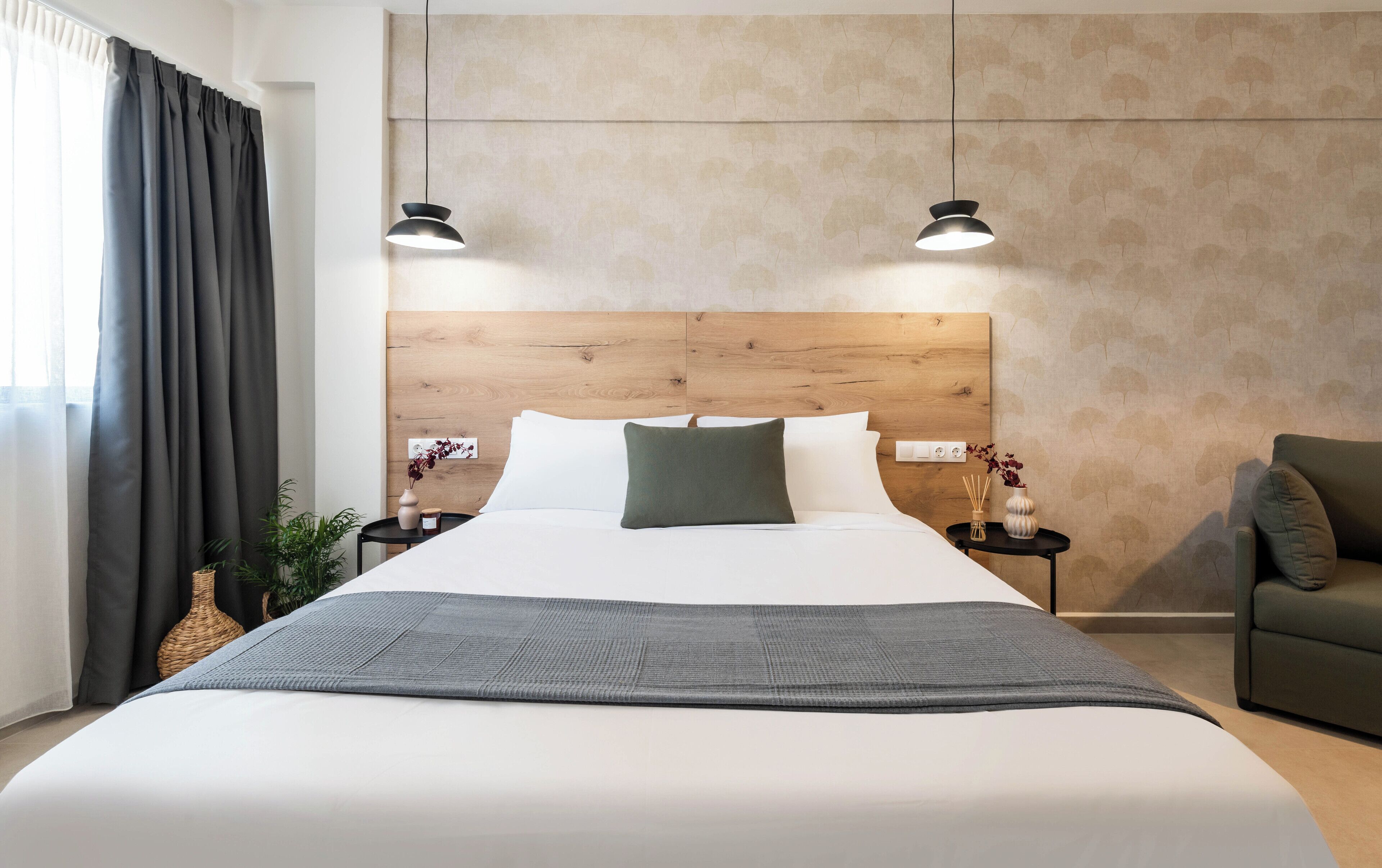 A modern hotel room with a wooden headboard, a large bed with white linens, two bedside lamps, a small side table with a vase, and a green armchair.