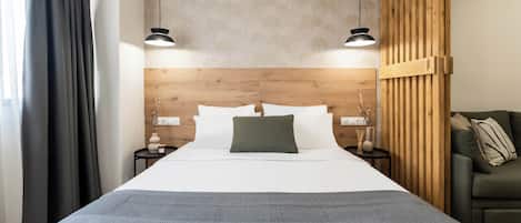 A modern hotel room with a large bed, wooden headboard, two bedside tables with lamps, a green sofa, and a view of the city.