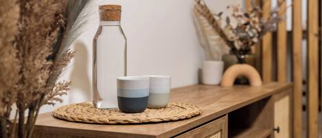 A wooden console table with a woven placemat, two ceramic cups, and a glass bottle with a cork stopper.