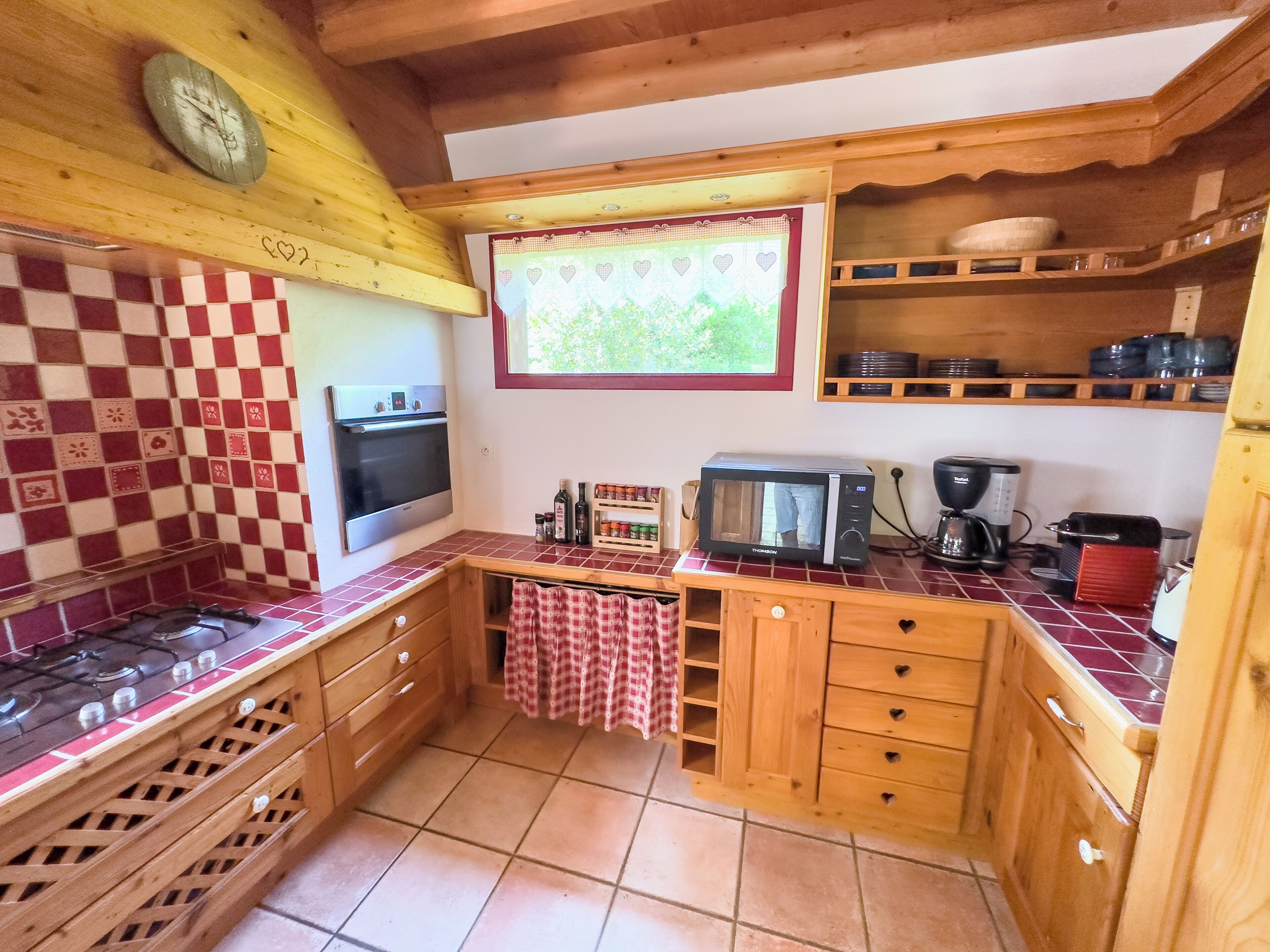 Una cucina con mobili in legno, zoccolo a scacchi, forno a microonde, fornello e finestra con vista sul verde.