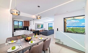 A modern kitchen with a large island, dining area with a table set for two, and a living area with a sofa and a large window overlooking a scenic view. - Major Villa Private Pool (Paralia Kourna)