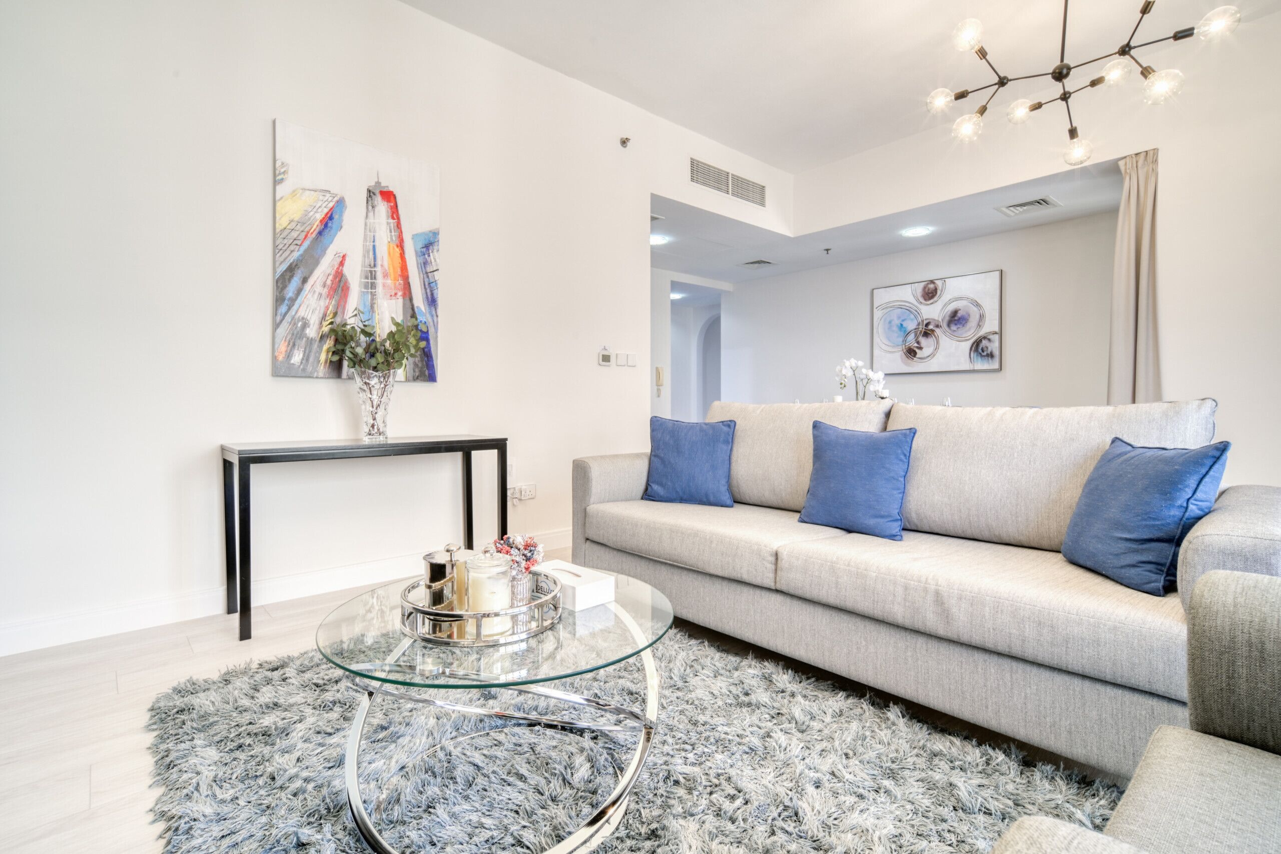 A modern living room with a glass coffee table, a grey sofa with blue cushions, and a cityscape painting on the wall.