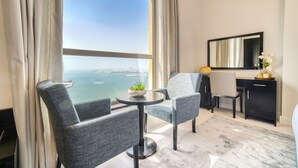 Ein modernes Hotelzimmer mit großem Fenster und Meerblick, zwei graue Polsterstühle um einen kleinen runden Tisch mit einer Pflanze, ein Schreibtisch mit Spiegel und eine schwarze Kommode.